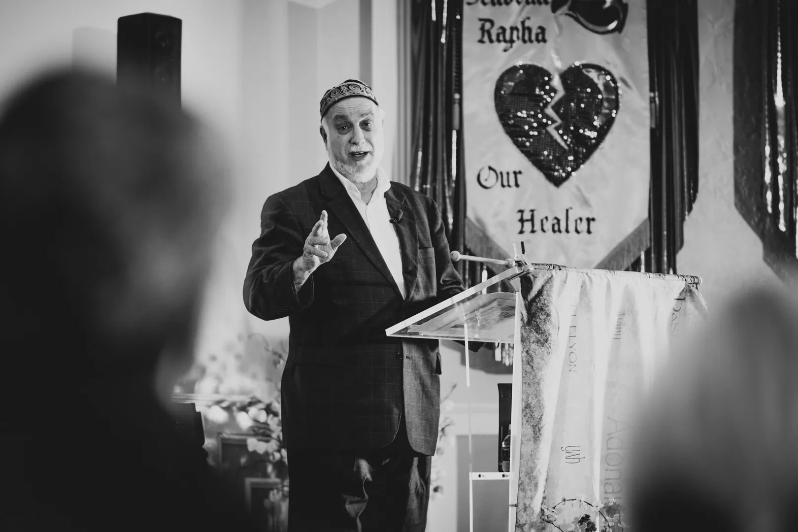 An elder in tallit and kippah leading the service beneath the congregation's Rapha banner.