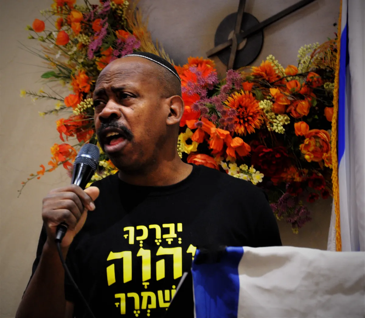 A worship leader singing into a microphone beside an arrangement of autumn flowers.
